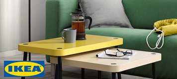 Un salon moderne avec un canapé vert et une table basse gigogne IKEA composée de deux pièces. La table supérieure est jaune vif, tandis que la table inférieure est en bois clair. Les deux tables ont des pieds noirs fins et un design minimaliste. Sur la table, on trouve une tasse, une cafetière en verre et des lunettes, ajoutant une touche fonctionnelle et conviviale à l'espace. À l'arrière-plan, un lampadaire blanc, un sac jaune et des rideaux gris complètent la décoration simple et chaleureuse.