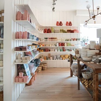 Intérieur d'une boutique de décoration avec des étagères blanches remplies de vases, bols colorés et accessoires, ainsi qu'une table en bois rustique exposant des articles de maison