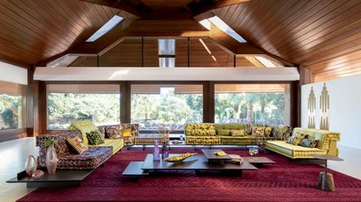 Canapé Mah Jong - Jean Paul Gaultier de Roche Bobois avec tissus floraux et colorés, disposé sur des plateformes noires, dans un salon lumineux au plafond en bois et grandes fenêtres avec vue sur la nature.