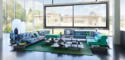 Canapé Mah Jong - Kenzo Takada UMI de Roche Bobois avec tissus bleus et verts à motifs floraux et tropicaux, disposé sur des plateformes noires, dans un salon moderne avec une vue sur un château à travers de grandes fenêtres.