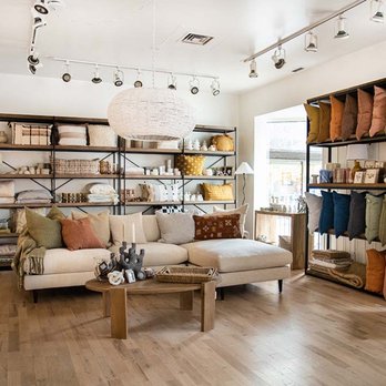 Salon d'exposition dans la boutique de décoration VdeV, avec un canapé beige décoré de coussins colorés, une table basse en bois et des étagères présentant des textiles et accessoires pour la maison.