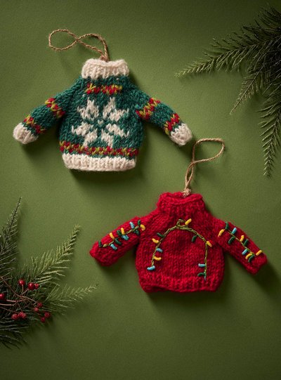 Deux décorations en forme de chandails tricotés accrochées sur un fond vert. L'un est vert avec un motif de flocon de neige blanc et des bordures colorées, l'autre est rouge avec des motifs de guirlandes lumineuses multicolores. Des branches de sapin et des baies rouges décorent les coins de l'image.