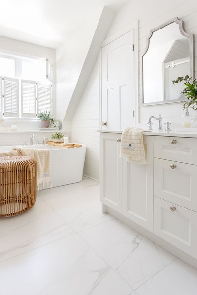 Salle de bain lumineuse avec meuble-lavabo blanc, robinetterie argentée, miroir décoratif, baignoire autoportante sous une fenêtre avec volets blancs, tabouret en rotin et sol en carrelage blanc effet marbre.