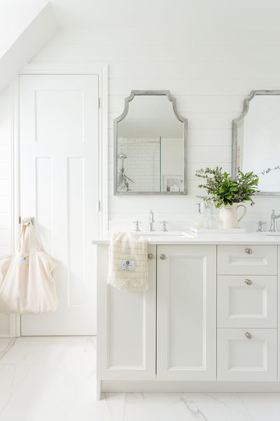 Salle de bain lumineuse avec meuble-lavabo blanc, robinetterie argentée, miroirs décoratifs gris, serviette beige, sac suspendu sur la porte et vase avec feuillage vert sur le comptoir.