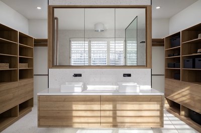 Salle de bain moderne avec meuble-lavabo double en bois naturel, vasques blanches rectangulaires, robinetterie noire murale, grand miroir encadré en bois, rangements ouverts en bois de chaque côté et fenêtres à persiennes blanches laissant entrer la lumière naturelle.