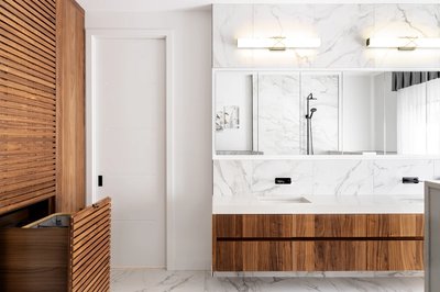 Salle de bain moderne avec murs en marbre blanc veiné de gris, meuble-lavabo suspendu en bois naturel, grand miroir, éclairage mural et douche minimaliste avec robinetterie noire. À gauche, meuble en bois à lattes et panier à linge intégré.