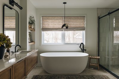 Salle de bain moderne avec baignoire autoportante blanche, robinet noir mat, meuble-lavabo en bois suspendu avec miroir arrondi, douche vitrée, étagères encastrées décorées et fenêtre avec stores en tissu beige.