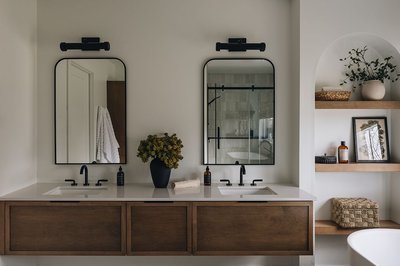 Salle de bain moderne avec meuble-lavabo double en bois, comptoir blanc, robinets noirs mats, miroirs rectangulaires arrondis avec luminaires noirs, étagères en bois encastrées avec accessoires décoratifs et vase noir avec fleurs séchées.