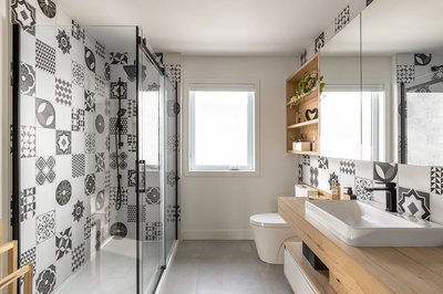 Salle de bain moderne avec carreaux à motifs noirs et blancs, douche vitrée à cadre noir, meuble-lavabo en bois clair avec vasque blanche, robinetterie noire, rangement mural en bois avec plantes décoratives, et fenêtre centrale laissant entrer la lumière naturelle.