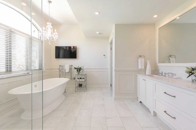 Salle de bain spacieuse et lumineuse avec baignoire autoportante, lustre en cristal, fenêtre cintrée avec stores, meuble-lavabo blanc, large miroir, téléviseur mural, table d’appoint chromée et sol en marbre blanc.