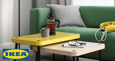 Un salon moderne avec un canapé vert et une table basse gigogne IKEA composée de deux pièces. La table supérieure est jaune vif, tandis que la table inférieure est en bois clair. Les deux tables ont des pieds noirs fins et un design minimaliste. Sur la table, on trouve une tasse, une cafetière en verre et des lunettes, ajoutant une touche fonctionnelle et conviviale à l'espace. À l'arrière-plan, un lampadaire blanc, un sac jaune et des rideaux gris complètent la décoration simple et chaleureuse.