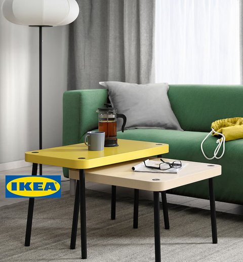 Un salon moderne avec un canapé vert et une table basse gigogne IKEA composée de deux pièces. La table supérieure est jaune vif, tandis que la table inférieure est en bois clair. Les deux tables ont des pieds noirs fins et un design minimaliste. Sur la table, on trouve une tasse, une cafetière en verre et des lunettes, ajoutant une touche fonctionnelle et conviviale à l'espace. À l'arrière-plan, un lampadaire blanc, un sac jaune et des rideaux gris complètent la décoration simple et chaleureuse.