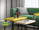 Un salon moderne avec un canapé vert et une table basse gigogne IKEA composée de deux pièces. La table supérieure est jaune vif, tandis que la table inférieure est en bois clair. Les deux tables ont des pieds noirs fins et un design minimaliste. Sur la table, on trouve une tasse, une cafetière en verre et des lunettes, ajoutant une touche fonctionnelle et conviviale à l'espace. À l'arrière-plan, un lampadaire blanc, un sac jaune et des rideaux gris complètent la décoration simple et chaleureuse.