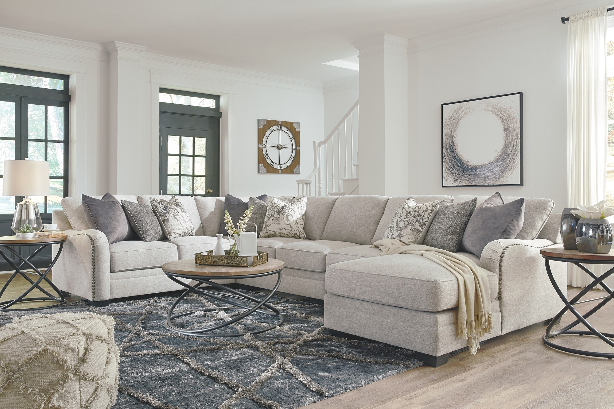 Salon moderne avec un canapé sectionnel beige, table basse ronde en bois et métal, tapis texturé gris, et décor élégant comprenant des coussins, une horloge murale et une œuvre d'art.