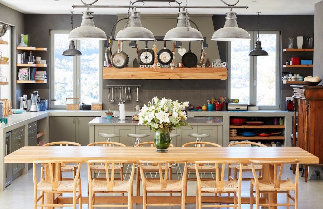 Cuisine lumineuse avec table en bois massif, chaises assorties, îlot central avec tabourets, étagères ouvertes et luminaires industriels suspendus.