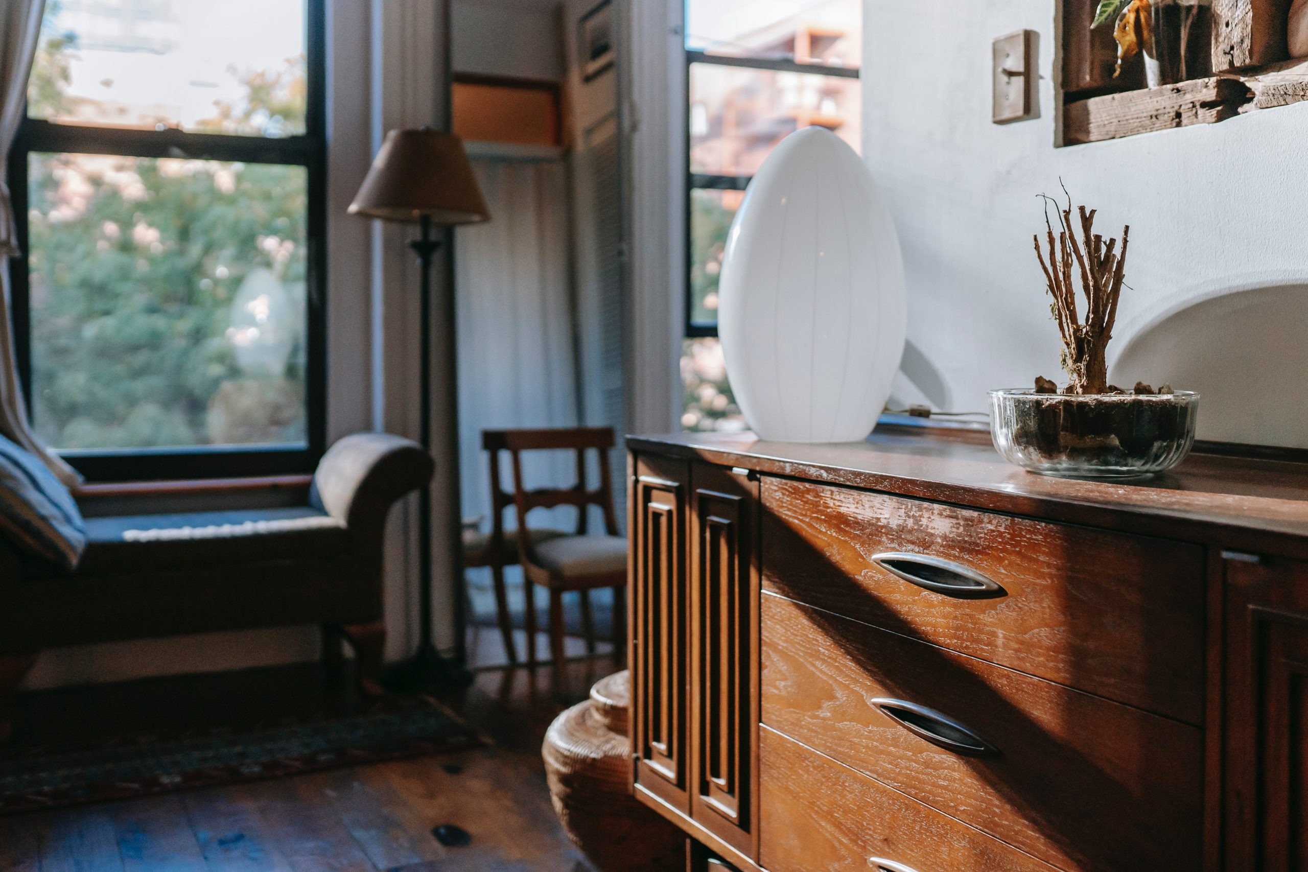 Buffet en bois vintage avec une lampe décorative blanche et une plante, dans un salon lumineux au style rétro avec vue sur l'extérieur de Chez Meublétout