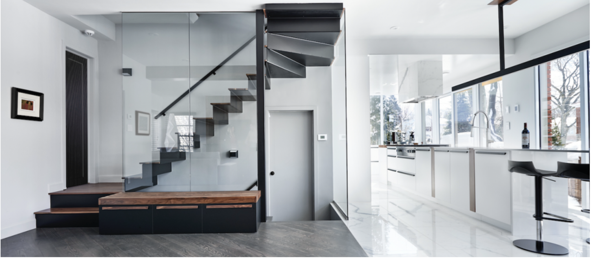 Escalier contemporain en métal noir et marches en bois, entouré de parois en verre, dans une maison lumineuse avec une cuisine ouverte et un design minimaliste. Lien vers site web de Ferland | Drouin